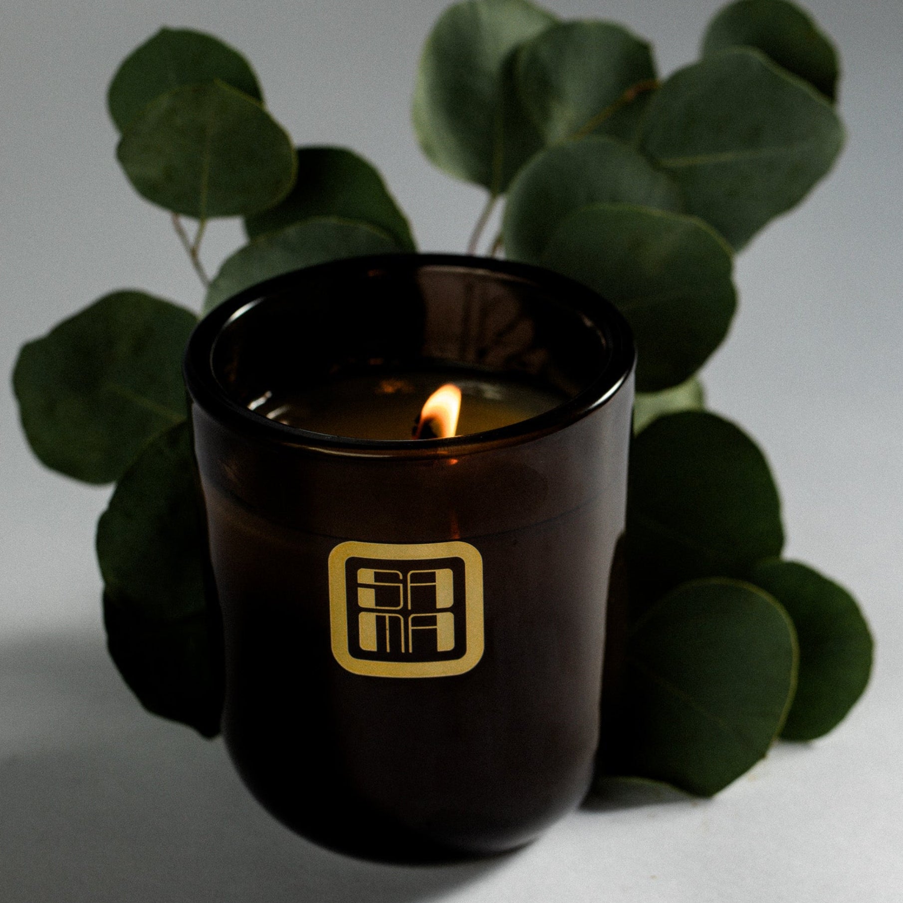 Brown candle with a gold logo surrounded by green leaves on a light background