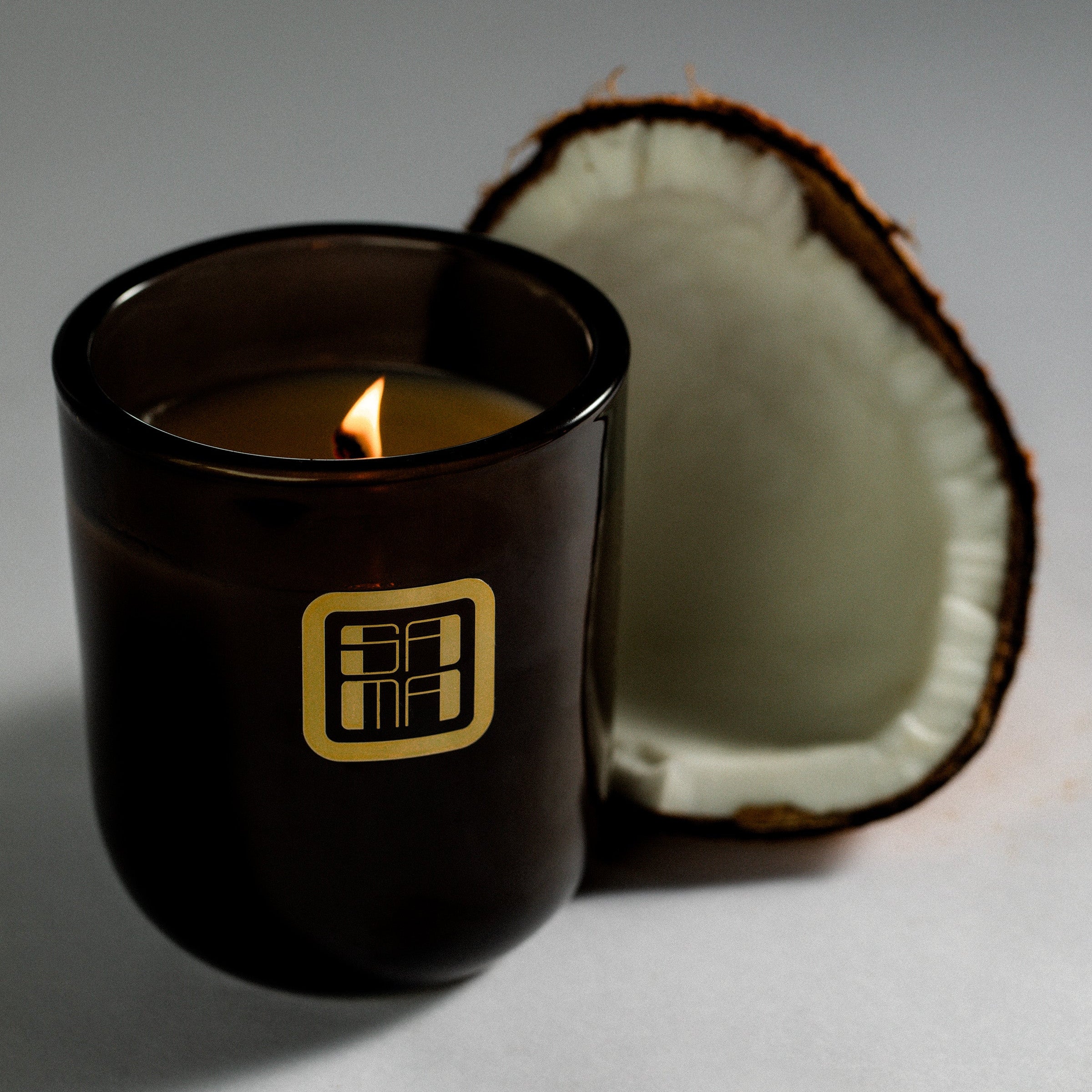 Black candle in a jar with a lit wick next to a halved coconut on a gray background