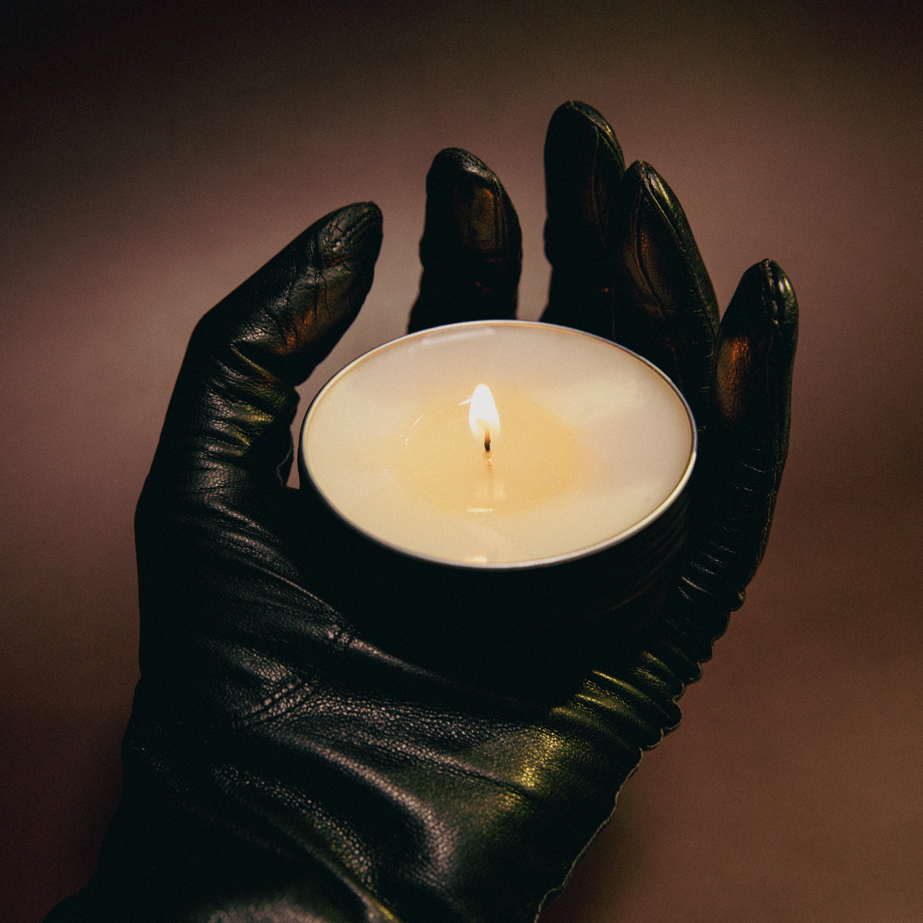 Hand wearing a black glove holding a lit candle against a dark background