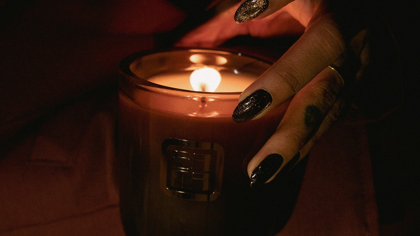 Hand holding a lit candle with dark nail polish in a dimly lit setting