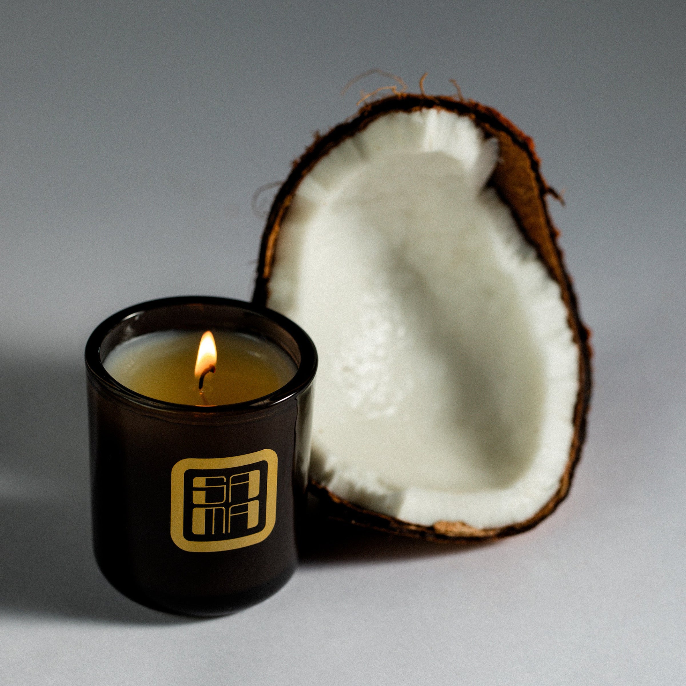 Candle in a black container with a gold logo next to a halved coconut on a gray background