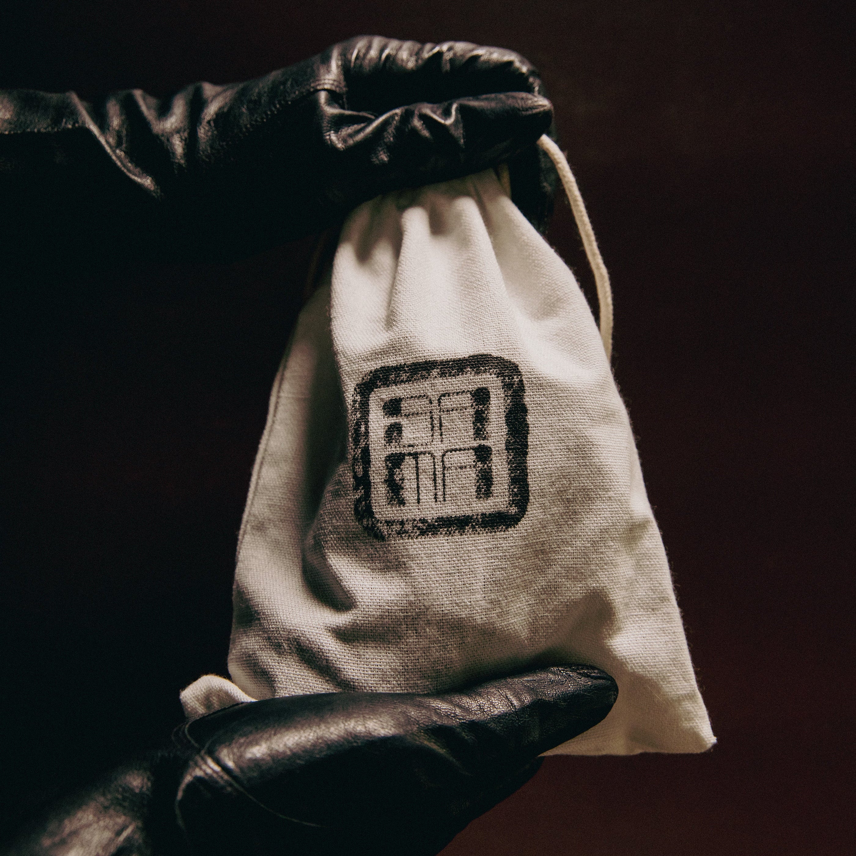 White drawstring bag with a black logo held by a gloved hand against a dark background